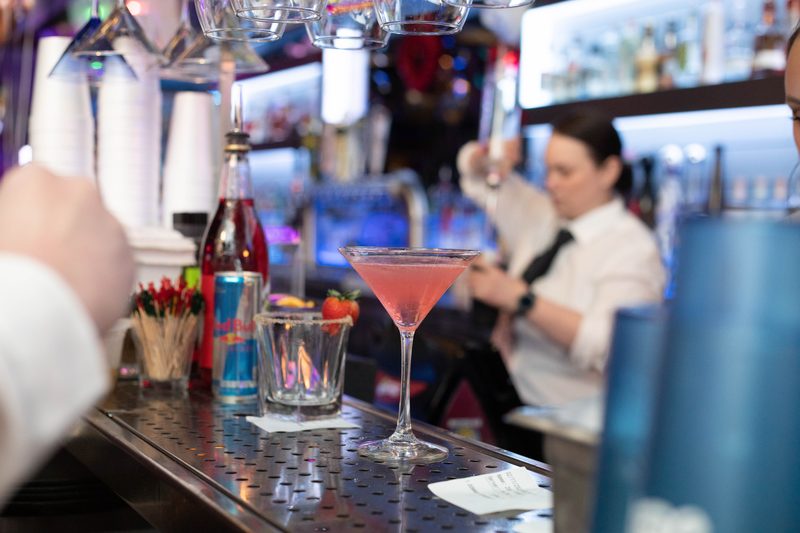 A pink cocktail in a martini glass on the bar with a bartender mixing behind it